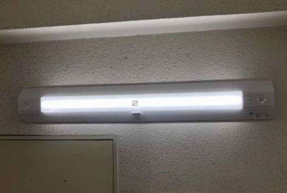 壁面の上部に取り付けられた直管型照明器具が点灯している様子。玄関や共用部の補助照明として使われている構図。