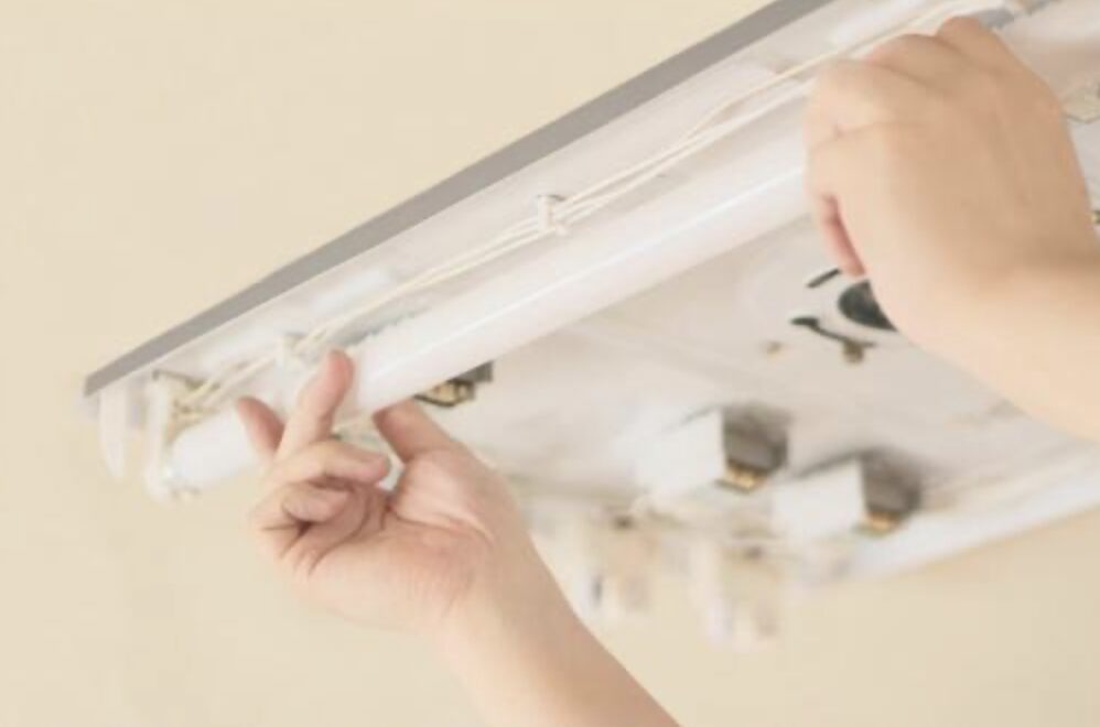天井に設置された直管蛍光灯器具の内部を、両手で蛍光灯を取り外し・取り付けしている作業中の様子。
