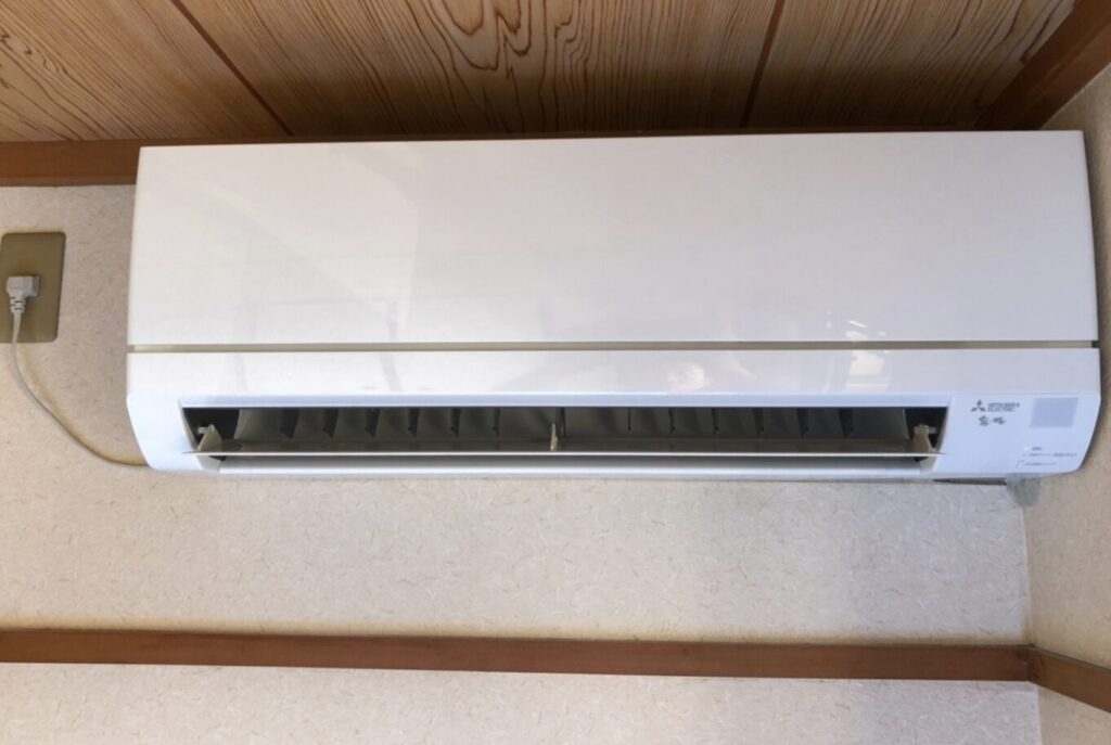 木目の天井下に設置された壁掛けエアコンを正面から撮影し、吹き出し口が開いている状態がわかる構図。