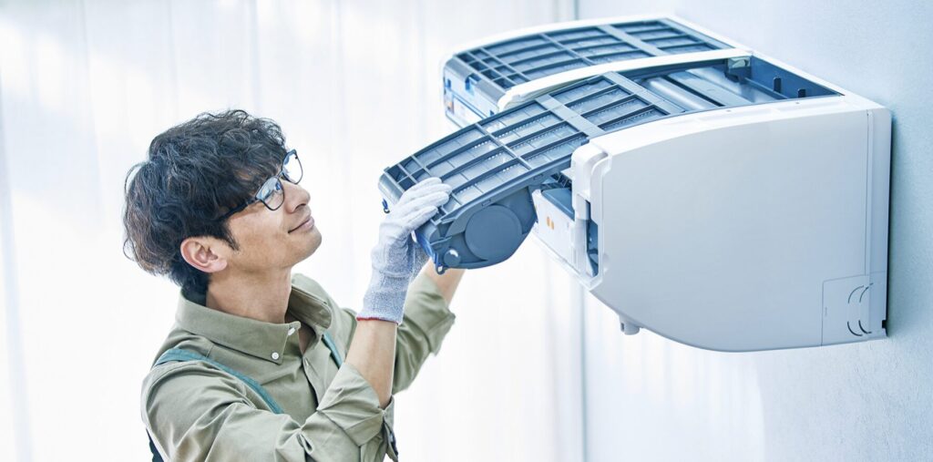 壁掛けエアコンの前面パネルを開け、手袋を着けた作業員がフィルターを取り外して点検している様子。