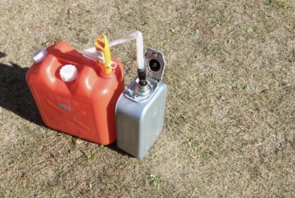 屋外の芝生の上に置かれた赤色の灯油用ポリタンクと金属製ポリ缶。ポリタンクからホースを使ってポリ缶へ灯油を移している様子が写っている。