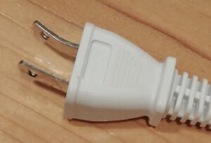 木目の床の上に置かれた白色の電源プラグ。2本の金属ピンのうち一方が外側に曲がっており、変形している状態がはっきり確認できる。