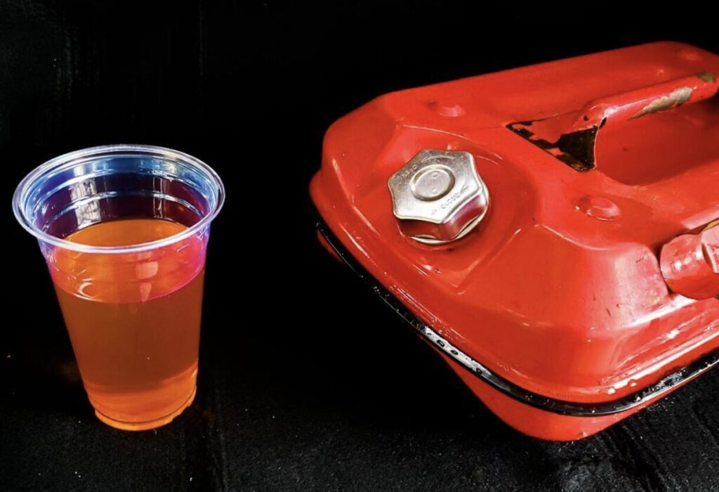 透明カップに入ったガソリンと、横に置かれた赤色の携行缶が並べられ、燃料の取り扱いリスクを示している構図。