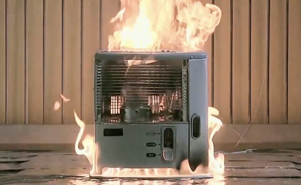 ガソリン誤給油により石油ストーブ上部から大きな炎が噴き出している実験映像で、周囲にも火が回っている様子。