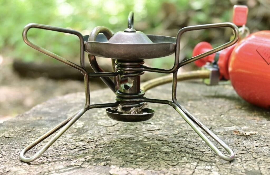 キャンプ用の液体燃料バーナーを地面に設置した状態で、脚部と燃焼ヘッドの構造が確認できるガソリンストーブの画像。