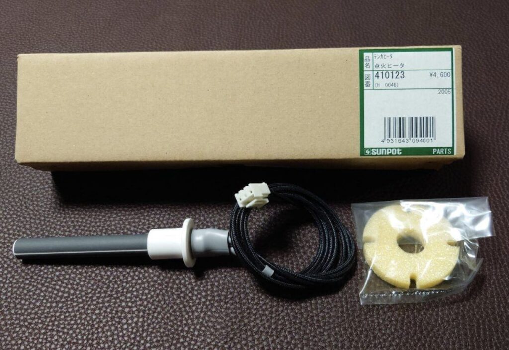 茶色の布地の上に置かれたサンポット製ストーブ用点火ヒーターの部品一式。品番やバーコードが記載された細長い箱の前に、配線付きの円筒状ヒーター部品と、袋入りの断熱材が並べられている。