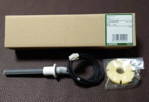 茶色の布地の上に置かれたサンポット製ストーブ用点火ヒーターの部品一式。品番やバーコードが記載された細長い箱の前に、配線付きの円筒状ヒーター部品と、袋入りの断熱材が並べられている。