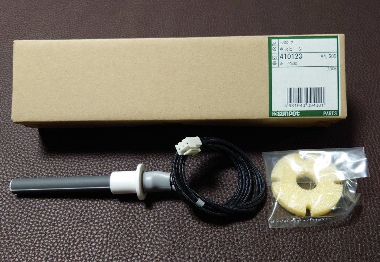 茶色の布地の上に置かれたサンポット製ストーブ用点火ヒーターの部品一式。品番やバーコードが記載された細長い箱の前に、配線付きの円筒状ヒーター部品と、袋入りの断熱材が並べられている。