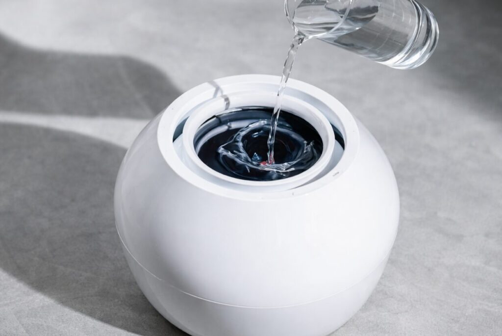 白い筐体の加湿器上部に、容器から水を注いで給水している様子。