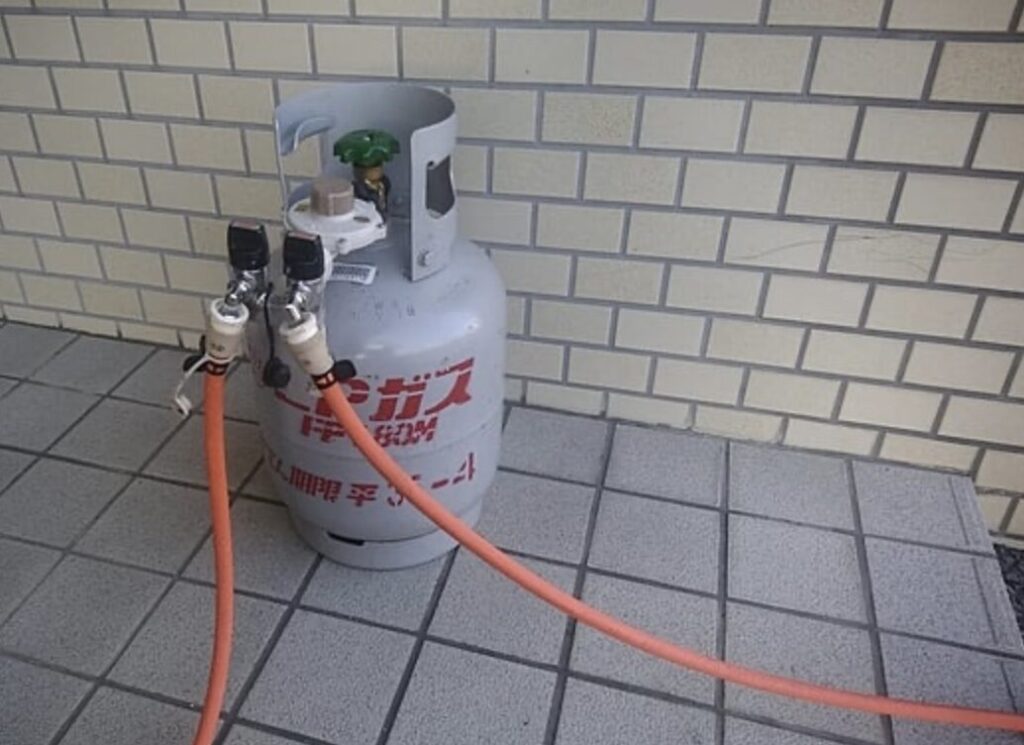 屋外の床面に設置されたLPガスボンベ1本と、分岐されたガスホースが接続されている様子。