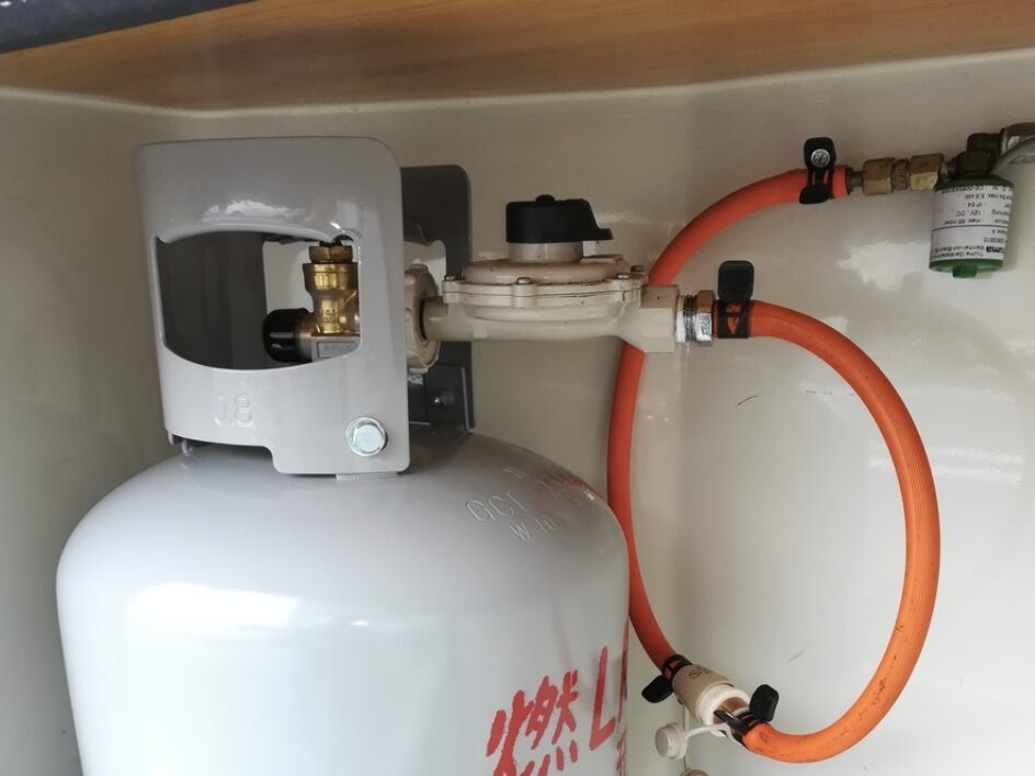 屋内設置されたLPガスボンベに調整器とオレンジ色のガスホースが接続されている状態。元栓と配管接続部が確認できる。