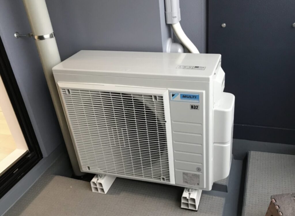 集合住宅のベランダに設置されたエアコン室外機。コンパクトな筐体で、配管スペースを抑えて設置されている様子。