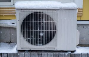 積雪した屋外に設置されたエアコン室外機。上部に雪が積もり、前面のファンと保護グリルが確認できる寒冷環境での設置状態。