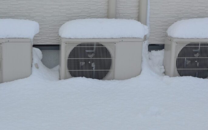 建物の外壁沿いに並べて設置された複数台のエアコン室外機が、下部まで雪に埋もれている状態。吸気不足が起きやすい設置環境を示している。
