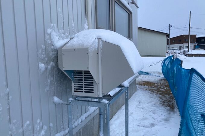 金属製の防雪カバーに覆われたエアコン室外機が屋外に設置されている様子。周囲は積雪が多く、冬季の運転環境を想定した構造が分かる。