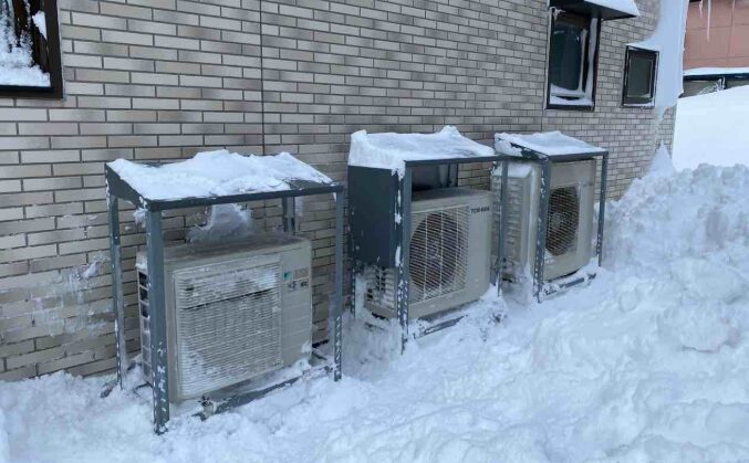 防雪フード付きのエアコン室外機が架台上に設置され、上部に積雪が残っている様子。降雪地域向けの対策が施された設置例。