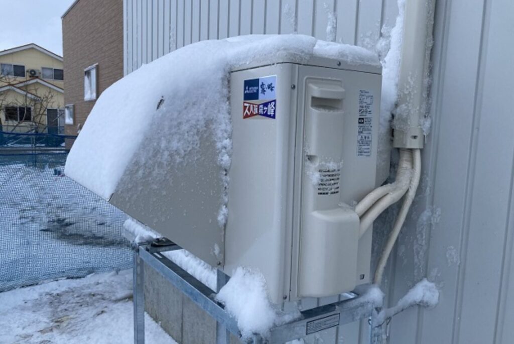 住宅外壁に架台設置された防雪フード付きエアコン室外機。上部に雪が積もり、配管や本体周囲にも着雪が見られる状態。