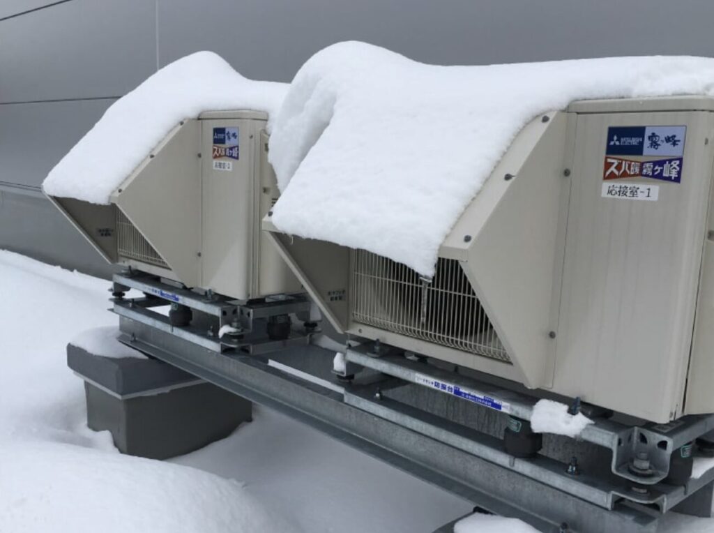 屋外に設置された防雪フード付きのエアコン室外機が2台並んでいる様子。フード上部に積雪があり、降雪地域での使用環境が分かる。
