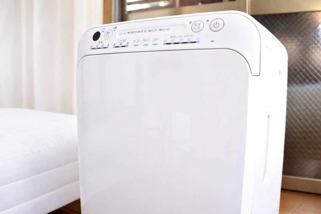 白い除湿機の操作パネルと前面パネルを斜め上から写した横長構図の写真。リビングの床に設置され、電源ボタンや湿度表示ランプが確認できる状態。
