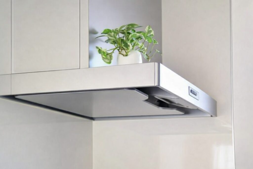 白いキッチンキャビネット下に設置されたスリム型レンジフードの写真。上部に観葉植物が置かれたシンプルな空間デザイン。