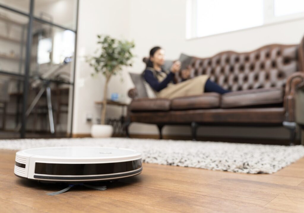 フローリングの床をロボット掃除機が走行し、奥のソファでは人物がくつろいでスマートフォンを操作しているリビングの様子。