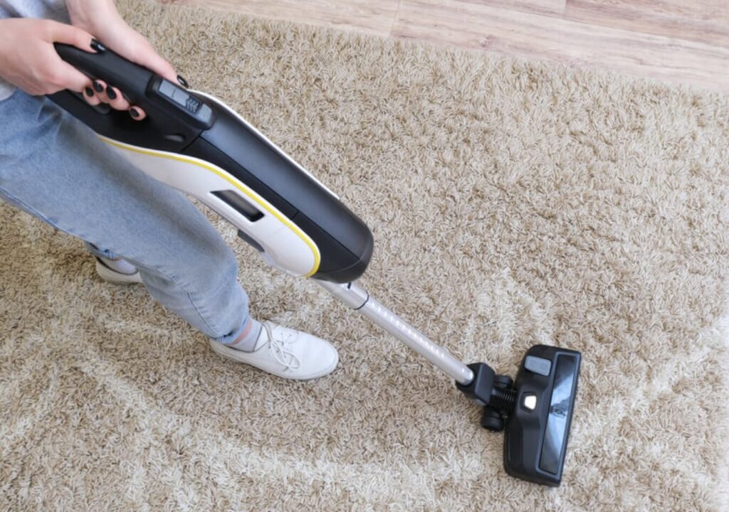 床のカーペットの上で、スティック型掃除機を手に持って掃除をしている様子。軽量タイプの掃除機を使った日常的な掃除シーン。