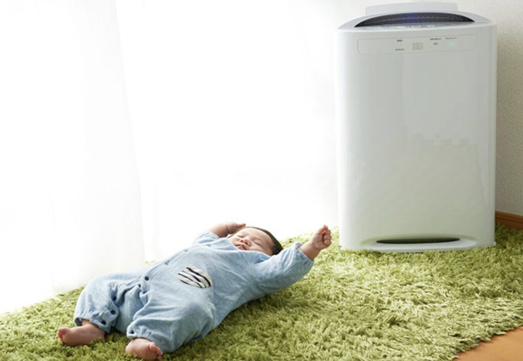 緑のラグの上で赤ちゃんが寝転び、その横で空気清浄機が稼働している室内の様子