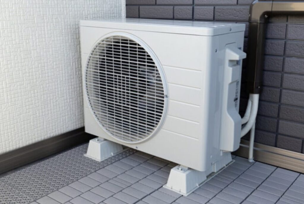 住宅の外壁横に設置されたエアコン室外機が、ブロックの上に固定されている様子