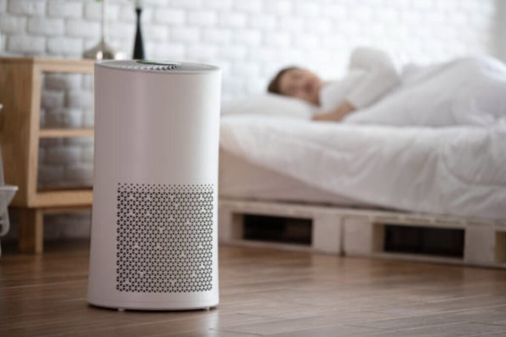 寝室の床に設置された空気清浄機。ベッドで休む人物のいる室内空気環境の改善イメージ。
