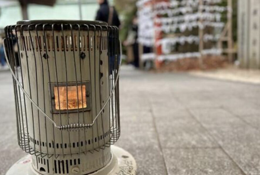 屋外の参道に設置された円筒型の石油ストーブ。金属製ガードの内側で炎が灯り、背景に人影と絵馬掛けがぼんやり写る様子。