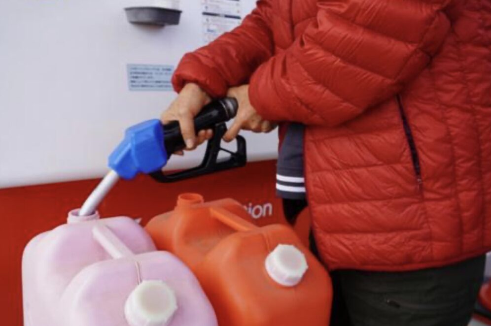 ガソリンスタンドで赤い灯油ポリタンクにノズルを差し込み給油している場面。冬用ダウンジャケットを着た人物の手元。