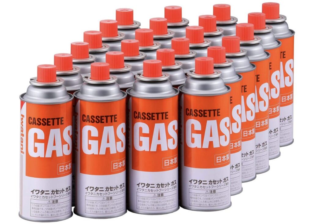 カセットガスボンベが複数本、赤いキャップ付きで整然と並べられている様子。日本製表示のあるオレンジ色ラベルのガス缶。