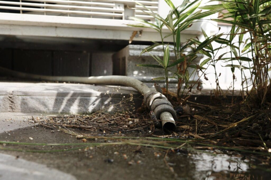 屋外に設置されたエアコンのドレンホース先端。周囲に落ち葉や泥が溜まり、水が排出されにくい状態