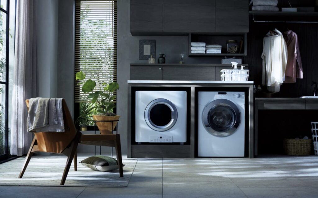 シックなダークトーンのランドリールームに並ぶドラム式洗濯機と乾燥機。観葉植物とブラインド窓がある落ち着いた室内空間。