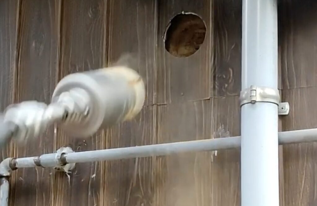 木製外壁に開けた丸穴をホールソーで加工している様子。屋外配管とともに排気ダクト施工中の作業シーン。