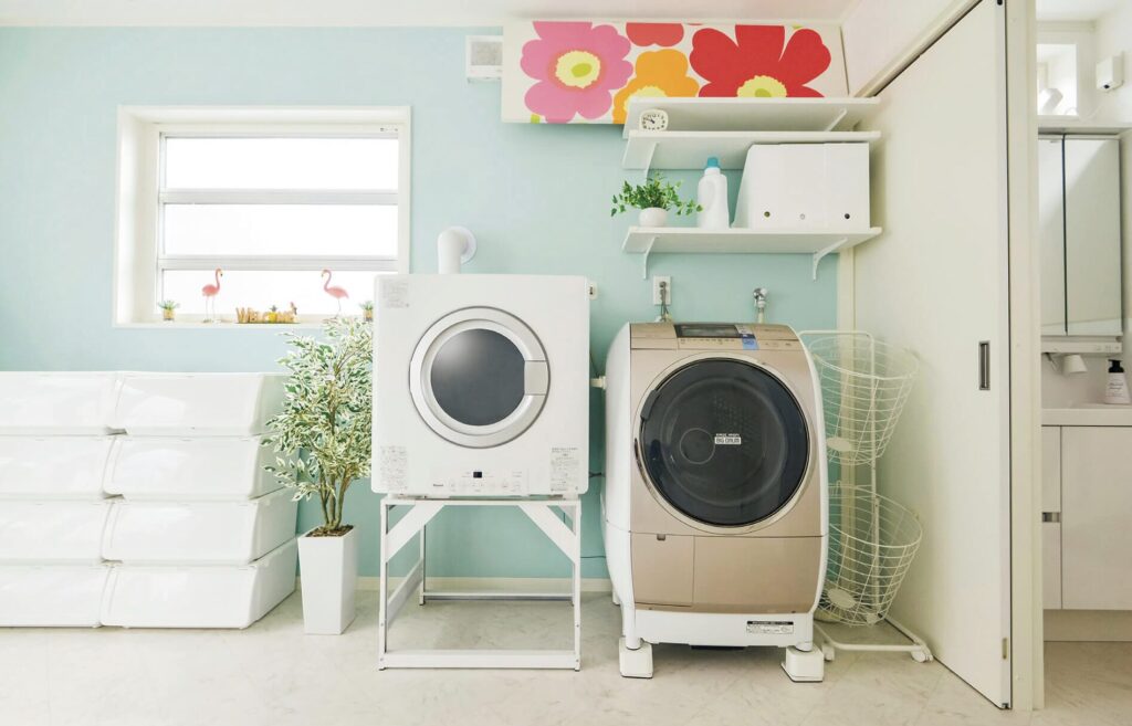 水色の壁と花柄アクセントクロスが特徴の明るいランドリールーム。スタンドに置かれたガス衣類乾燥機とドラム式洗濯機が並ぶ空間。
