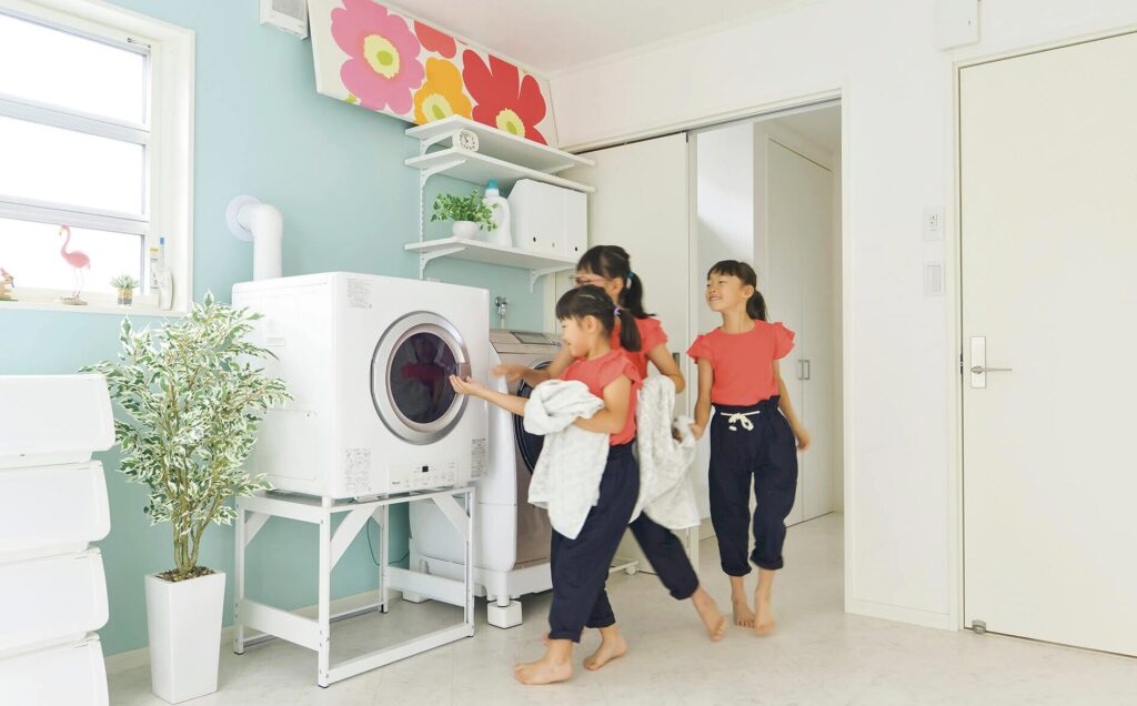 子どもたちがランドリールームでガス衣類乾燥機に洗濯物を入れようとしている明るい家庭の風景。
