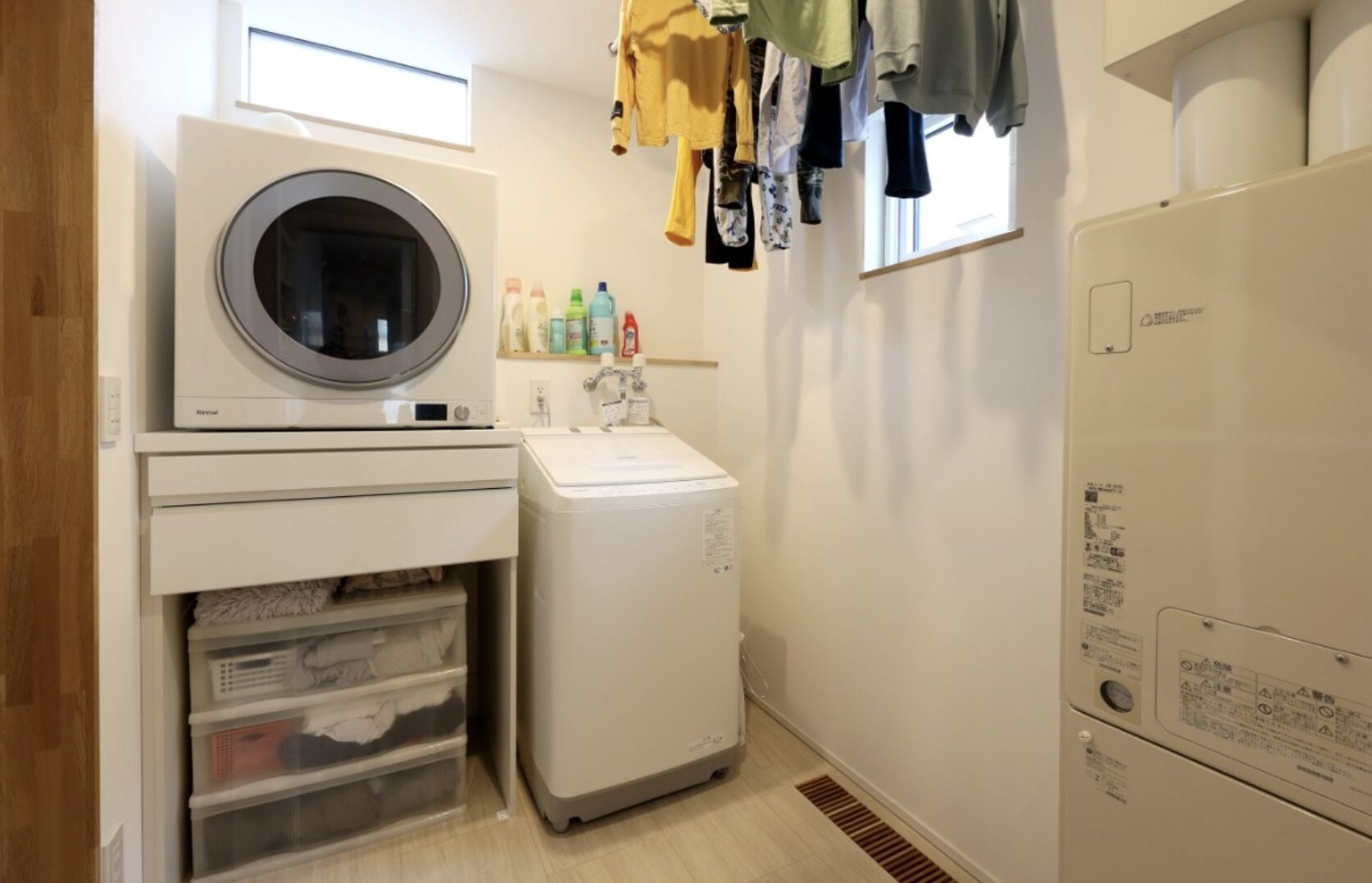 壁面に設置された白いガス衣類乾燥機と縦型洗濯機が並ぶコンパクトなランドリールーム。天井付近には室内物干しがあり、洗剤ボトルや収納ケースが整理されている様子。