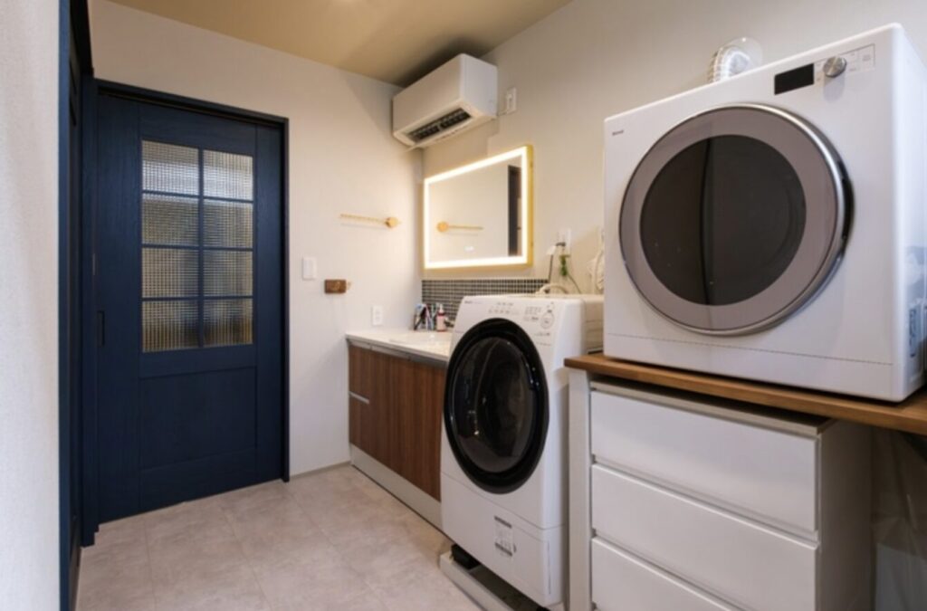 木目カウンターと間接照明付きミラーがある洗面室に設置されたガス衣類乾燥機とドラム式洗濯機。落ち着いた内装デザインのランドリースペース。