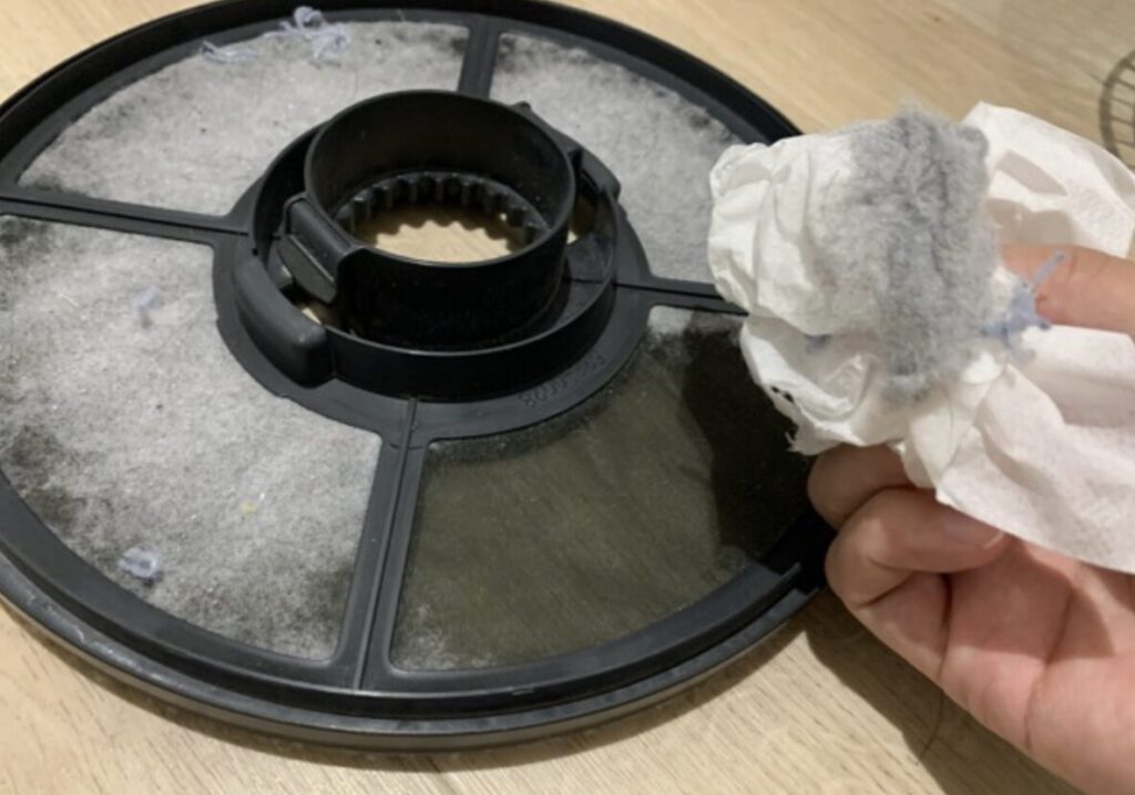 取り外した乾太くんの丸型フィルターに溜まった糸くずをティッシュで拭き取っている様子。掃除中の実写イメージ。