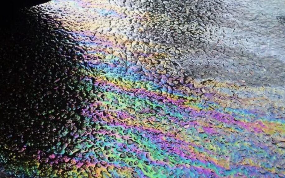 路面に広がった液体の表面に虹色の膜が浮かび上がっている様子。油分が混ざった水たまりの拡大イメージ。