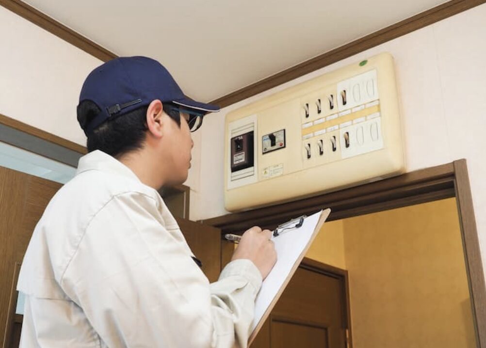 作業員がブレーカーを点検しながらクリップボードに記録している様子。室内ドア上部に設置された分電盤の前での確認作業。