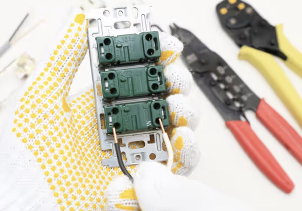 作業用手袋を着用し、コンセント裏側の端子に電線を接続している手元のアップ。周囲に圧着工具が置かれている。