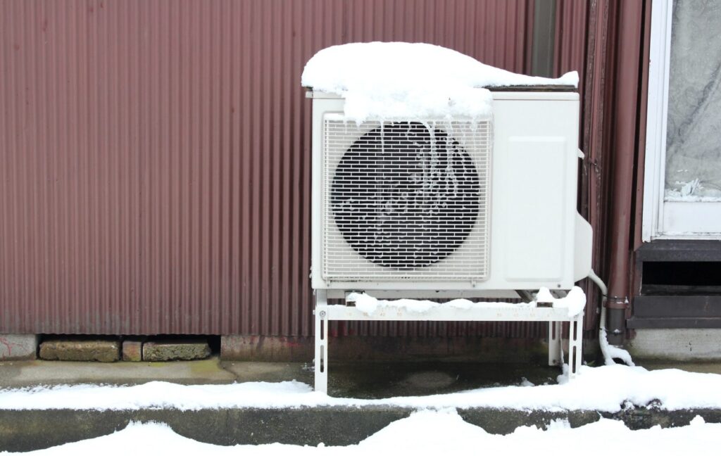 雪が積もった屋外に設置されたエアコン室外機。架台の上に設置され、周囲の地面や上部に積雪が見られる状況。