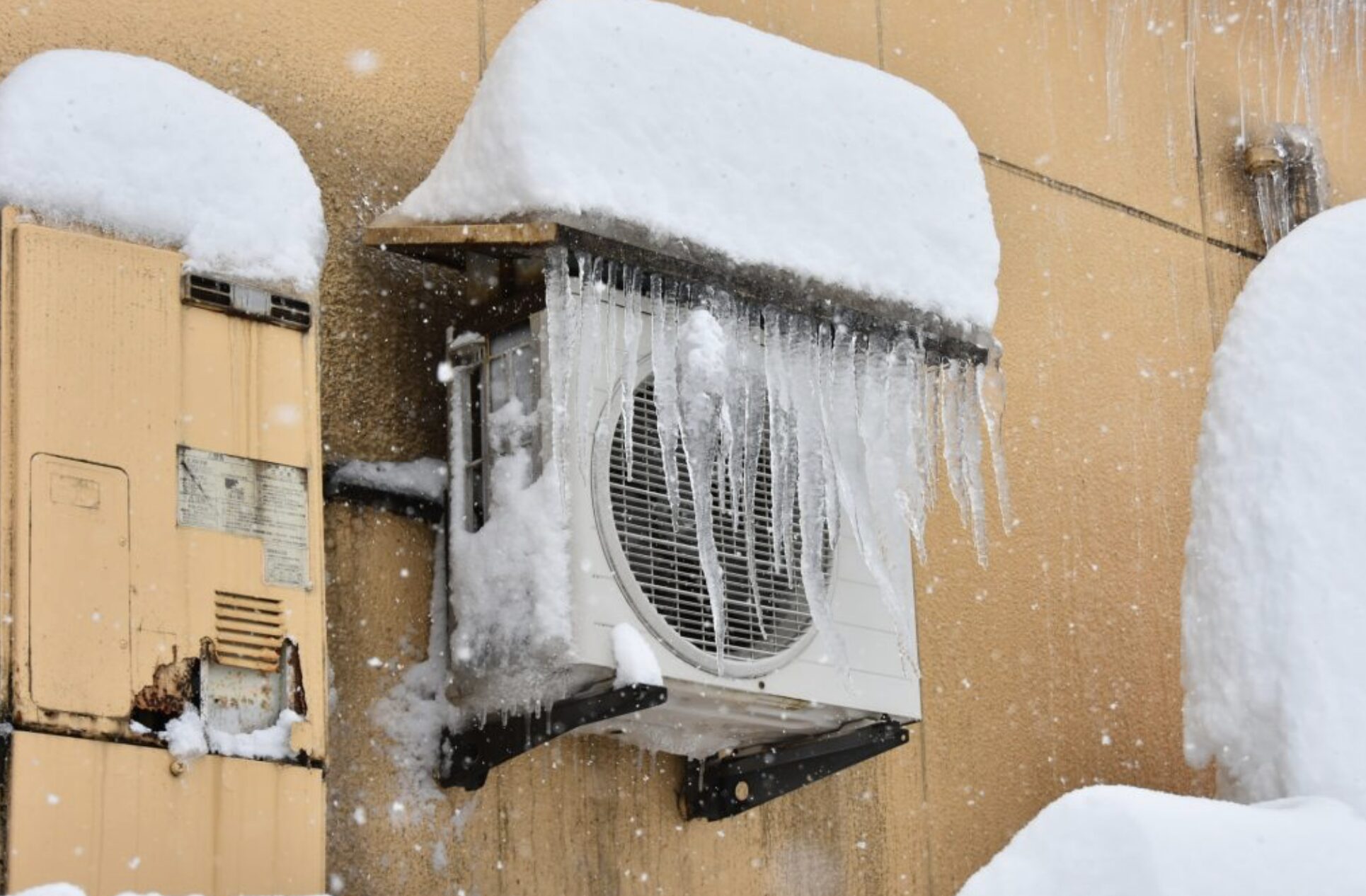 外壁に設置されたエアコン室外機に雪が積もり、上部から長いつららが垂れ下がっている様子。降雪中の寒冷環境での設置状況。