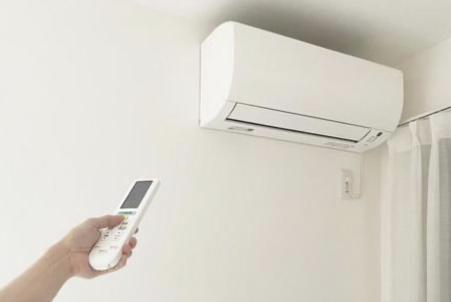白い壁に設置された壁掛けエアコンを、手に持ったリモコンで操作している様子。シンプルな室内空間で冷暖房の使用イメージを表している。