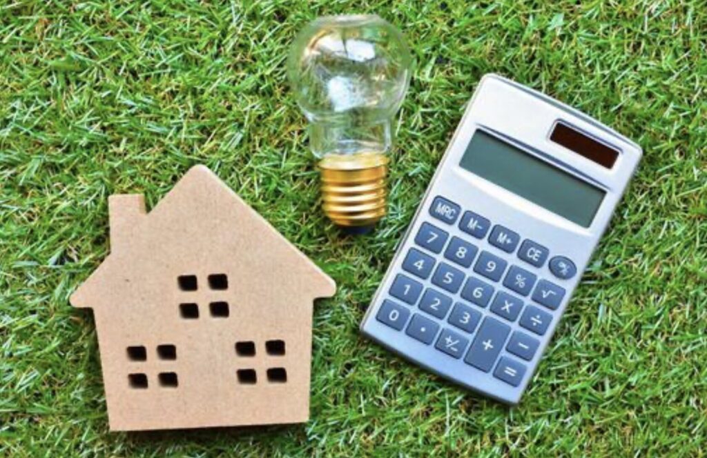 緑の芝生の上に、段ボール製の家型オブジェ、透明な電球、グレーの電卓が横一列に配置されたイメージ。住宅と光熱費の計算を連想させる構図。