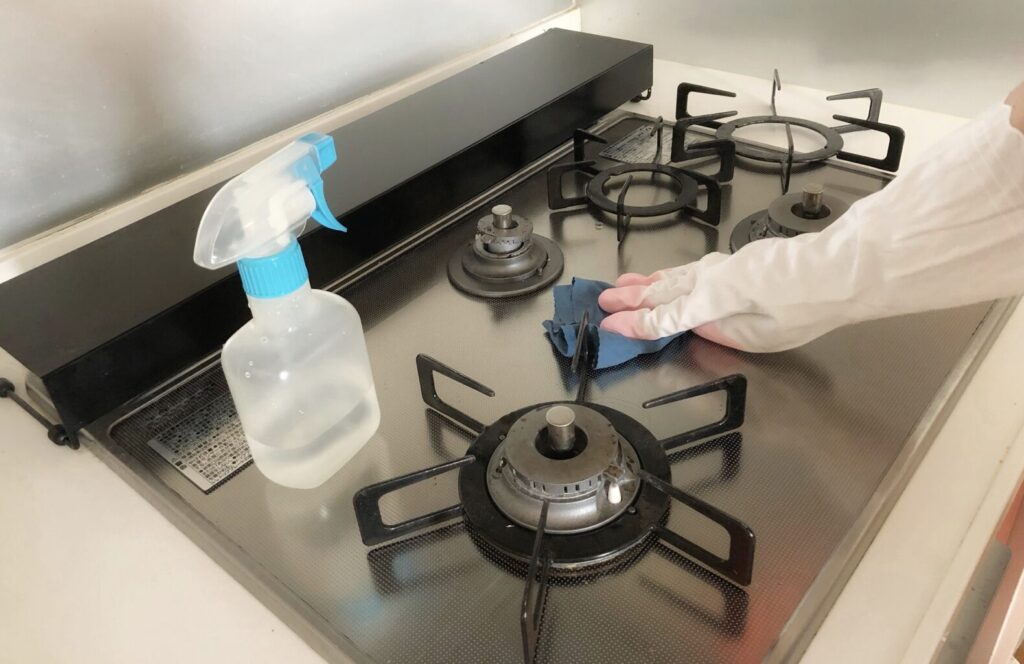ガスコンロの天板を布で拭き掃除している様子。スプレーボトルと手袋を着用した手が写っている。
