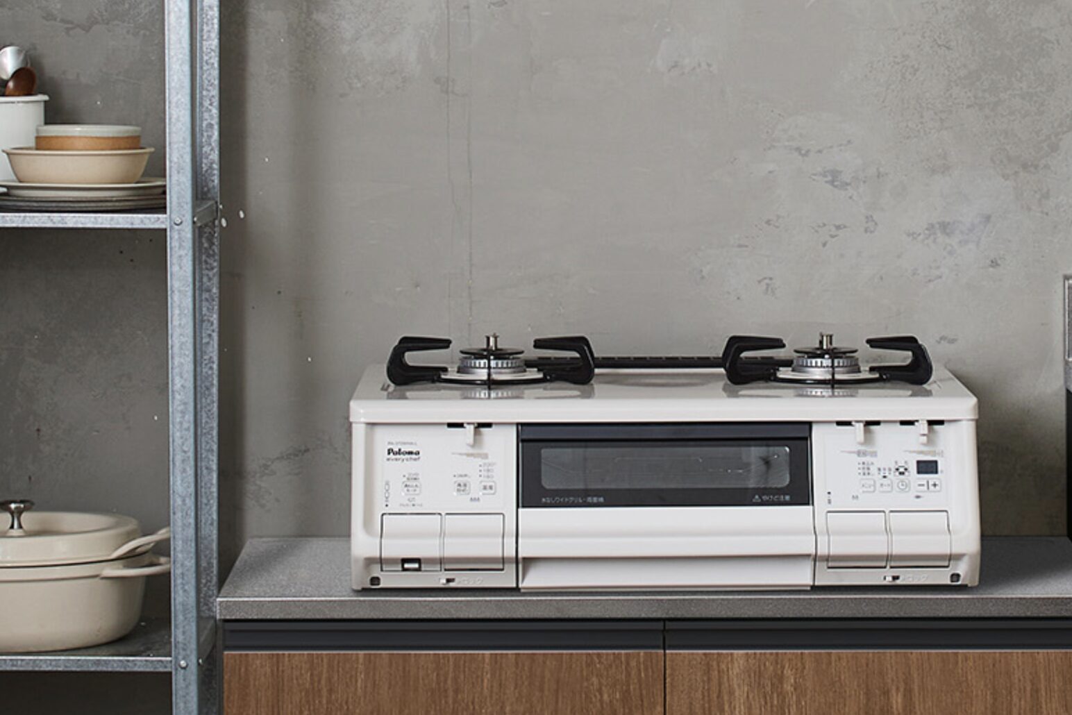 キッチンカウンターに設置された白色の据え置き型二口ガスコンロ。中央に魚焼きグリルを備え、背後はグレーの壁面、左側に金属ラックと調理器具が置かれている。