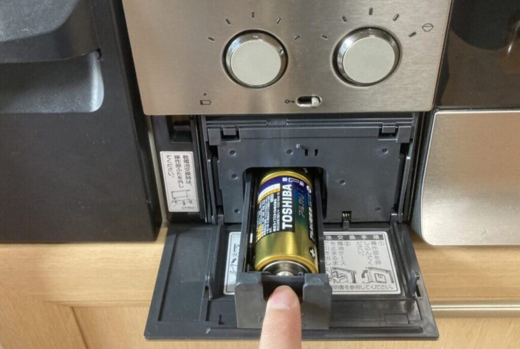 ガスコンロ前面の電池ボックスを開き、単1アルカリ乾電池を差し込んでいる手元の様子。